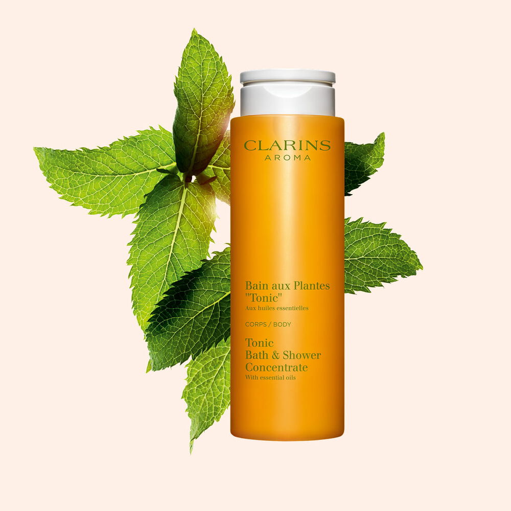 Kuva Clarins Tonic Bath and Shower Concentrate suihkugeelist&auml; sek&auml; mintun ja rosmariinin lehdist&auml; oranssia taustaa vasten.