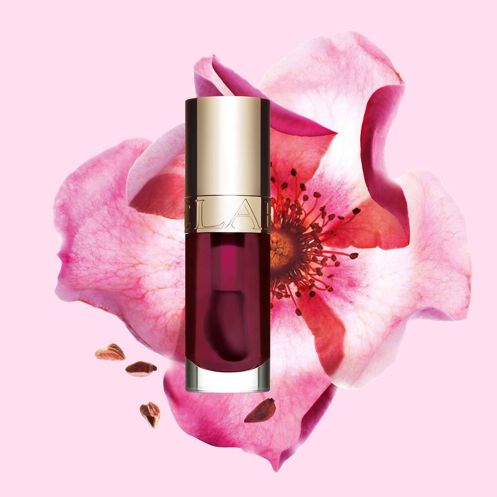 Clarins Lip Comfort Oil Fig packshot, asetettuna lähellä ruusunmarjaa vaaleanpunaisella taustalla.