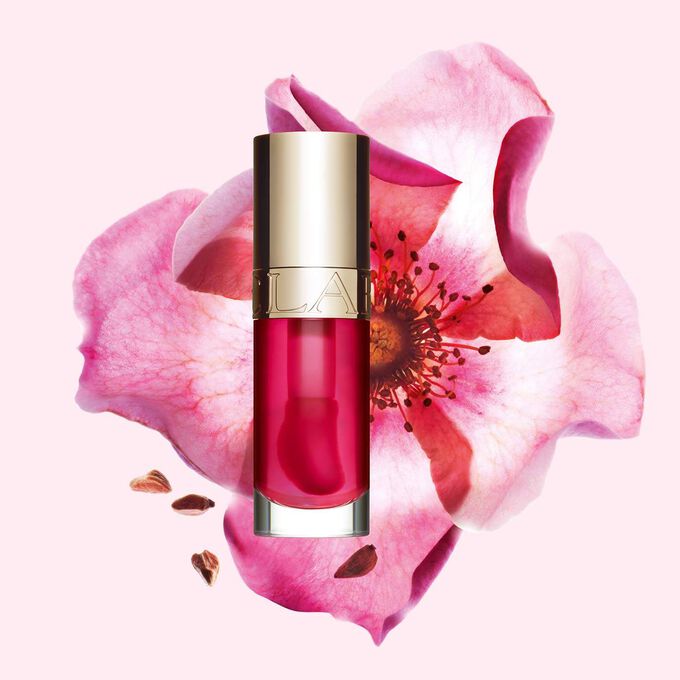 Packshot Clarins Lip Comfort Oil -tuotteesta Pitaya, asetettuna lähelle ruusunmarjaa vaaleanpunaiselle taustalle.
