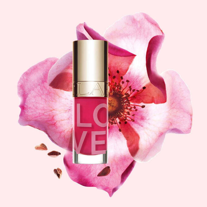 Tuotekuva Lip Oil Limited Edition Love Collection -huuli&ouml;ljyst&auml; ruusun edess&auml; vaaleanpunaisella taustalla, joka korostaa tuotteen elinvoimaista s&auml;vy&auml;.
