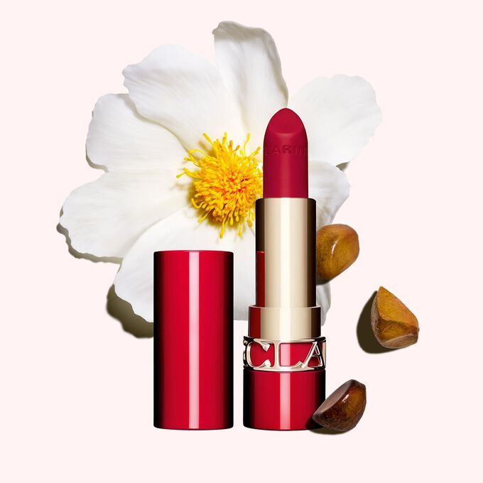 Packshot Clarins matta huulipuna Joli Rouge 742 V, kameliakukan ja sheansiemenen vieressä vaaleanpunaisella taustalla.