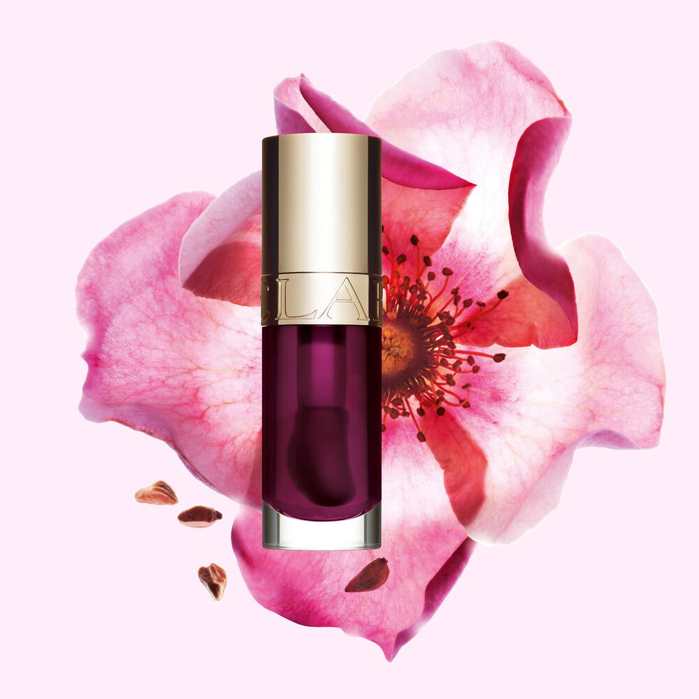 Packshot Clarins Lip Comfort Oil -tuotteesta s&auml;vyss&auml; Plum, sijoitettuna ruusunmarjakukan vierelle vaaleanpunaiselle taustalle.
