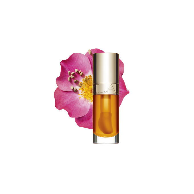 Packshot Clarins Lip Comfort Oil -tuotteen Honey-värissä, asetettuna lähelle ruusunmarjakukkaa vaalean persikkapohjan päälle.